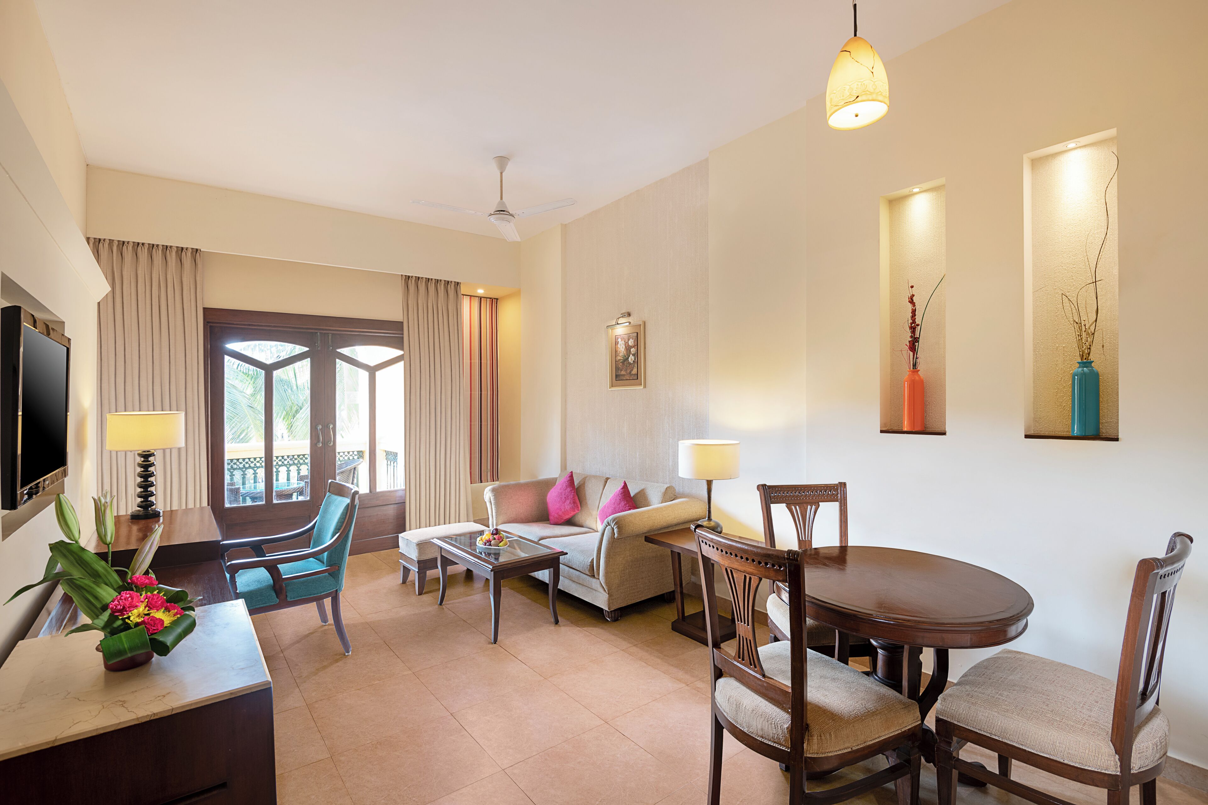 Habitación de hotel con sofá, sillas, mesa de comedor, televisor y escritorio con lámpara y flores.