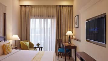 A hotel room with a large bed, two chairs, a desk with a lamp, a TV, and a window with curtains.