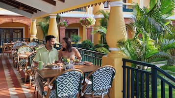 A restaurant with outdoor seating, featuring patterned chairs and tables, surrounded by greenery and architectural columns.