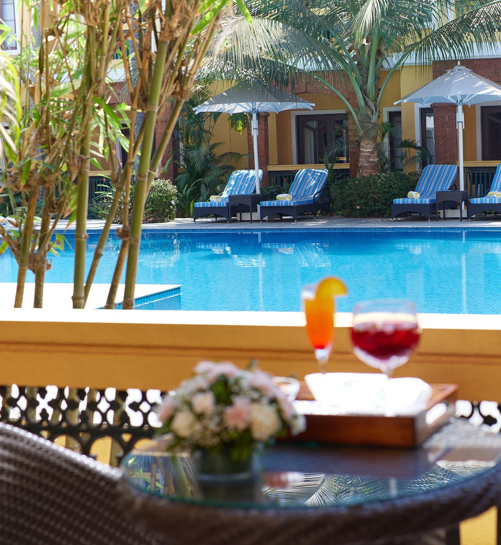 A poolside view with lounge chairs and umbrellas, a glass table with a drink and flowers, and a clear blue pool.