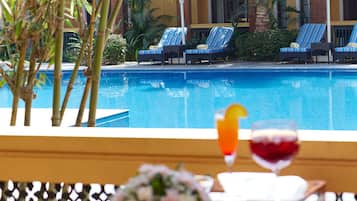 A poolside view with lounge chairs and umbrellas, a glass table with a drink and flowers, and a clear blue pool.