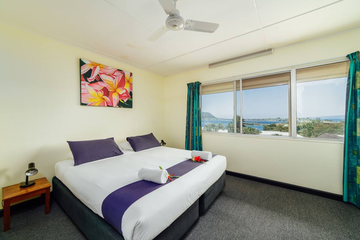 A hotel room with a bed, a ceiling fan, a window with curtains, and a painting on the wall.