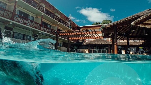 Una piscina con agua cristalina, un edificio con balcones y una zona techada con mesas y sillas.