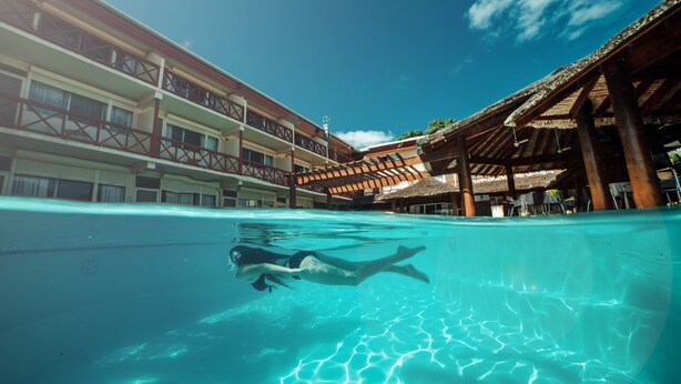 An indoor swimming pool with clear water, a person swimming, and a building with balconies and a thatched roof in the background.