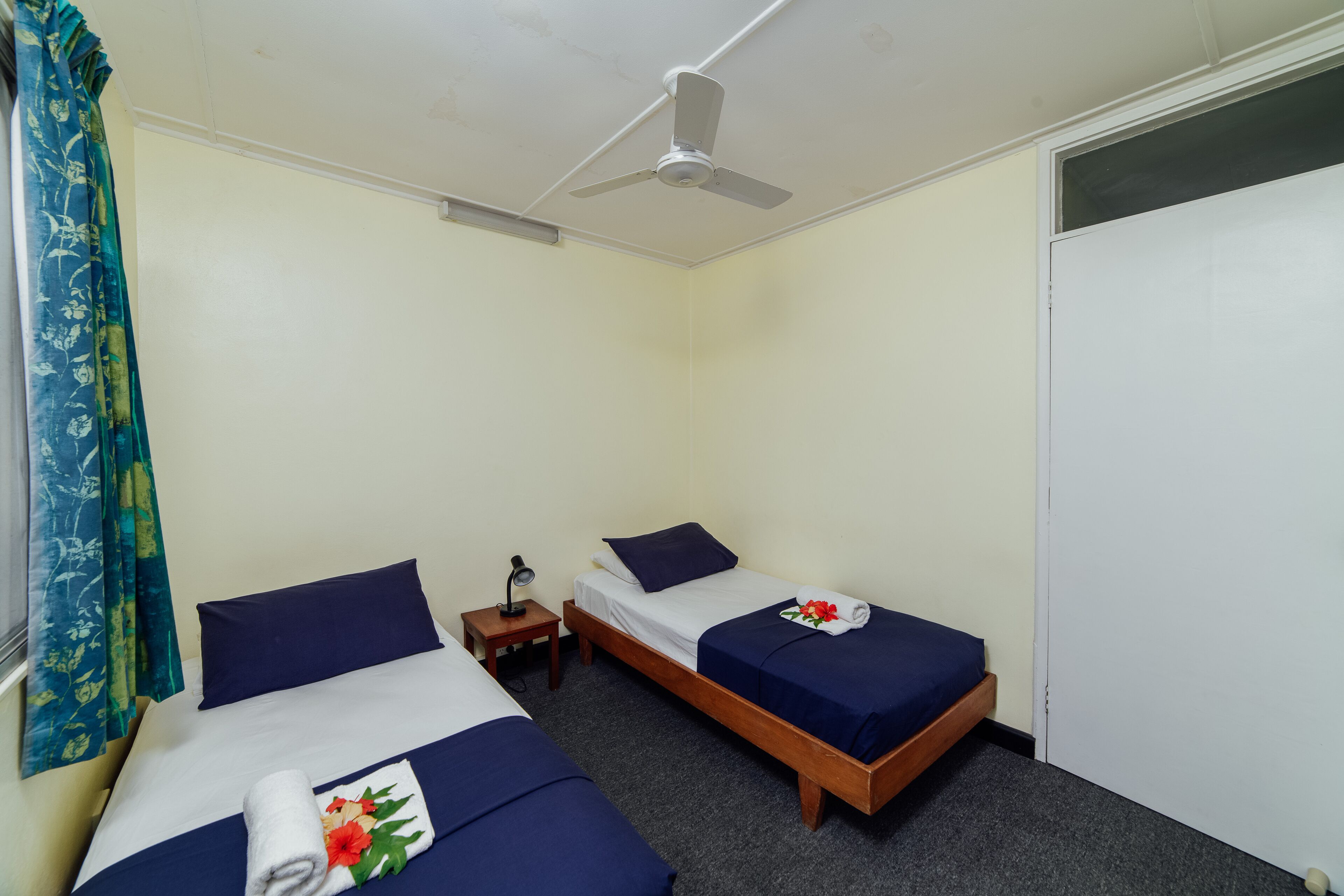 A room with two single beds, a ceiling fan, a small table with a lamp, and a window with curtains.