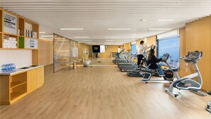 Ett gym med träningsutrustning, en reception och utsikt över staden genom stora fönster.