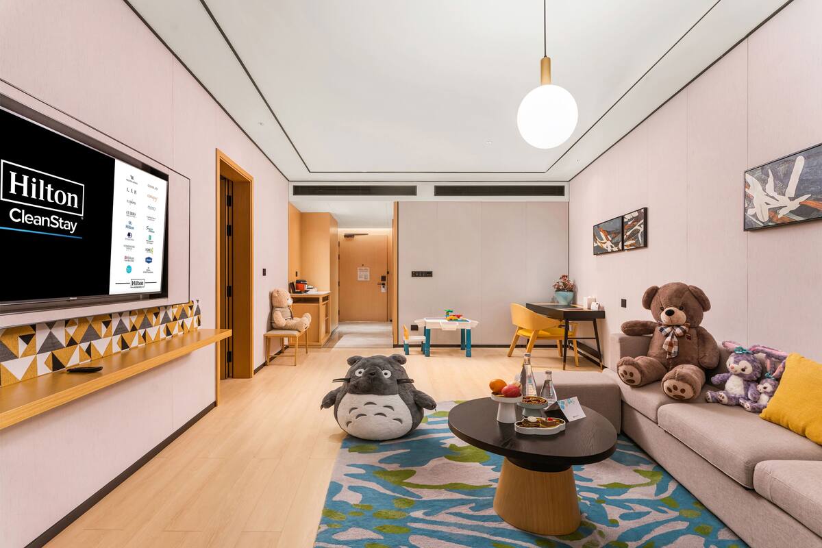 Une chambre d’hôtel moderne dotée d’un grand téléviseur affichant le logo Hilton CleanStay, d’un canapé, d’une table basse et d’un pouf en forme d’ours.