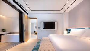 Ett modernt hotellrum med en stor säng, en platt-TV som är monterad på väggen, ett sängbord och ett badrum med badkar och handfat.