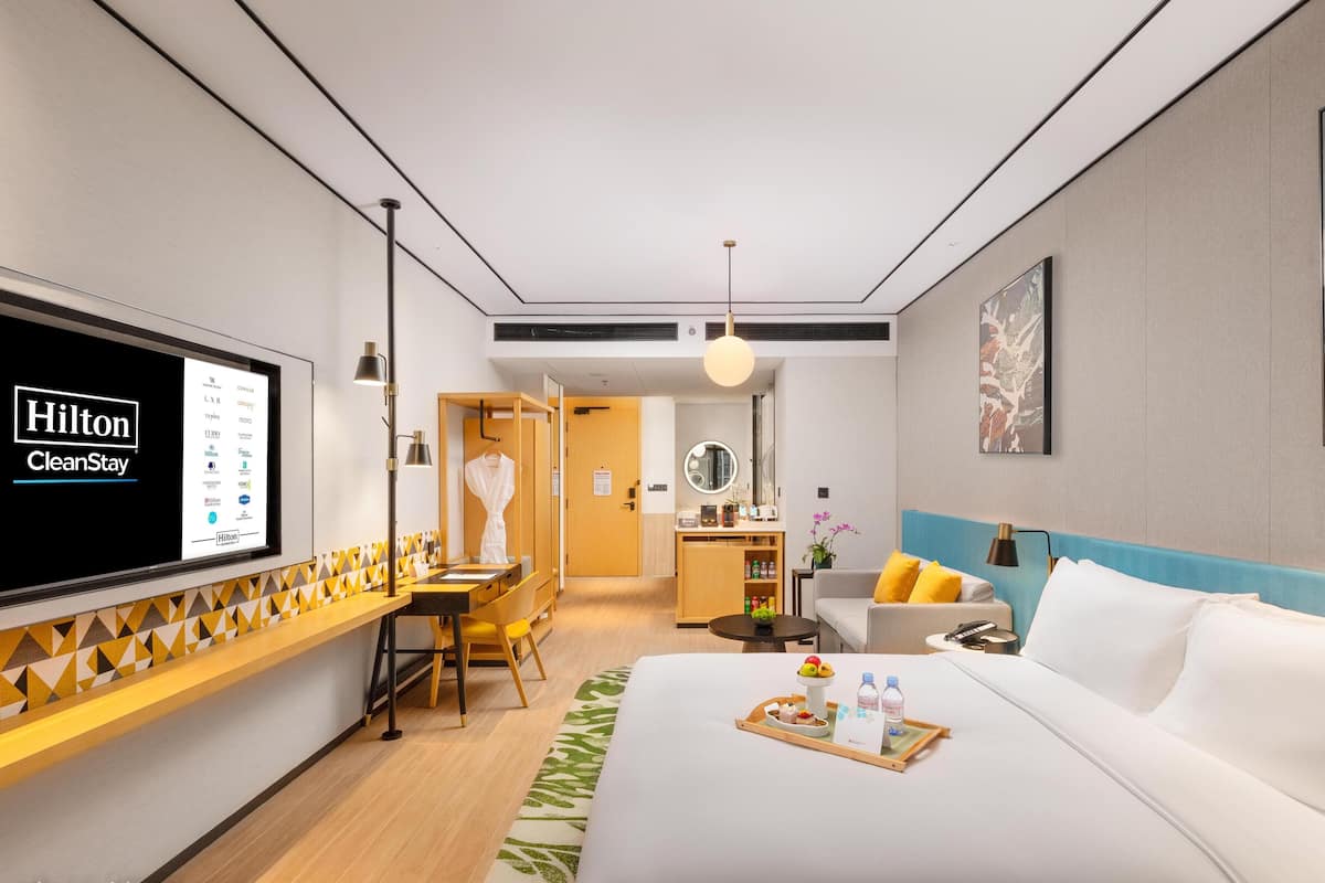 Une chambre d’hôtel moderne équipée d’un lit, d’un bureau, d’une chaise, d’une petite table, d’un miroir et d’un écran de télévision affichant le logo Hilton CleanStay.