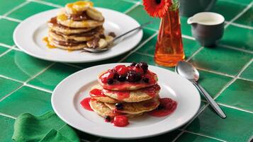 Ein Frühstückstisch mit Pfannkuchen und Beeren auf Tellern, einer Vase mit einer Blume und einem Teekännchen auf einer grün karierten Tischdecke.