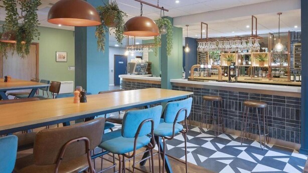 A restaurant with a bar area, blue chairs, and hanging plants. - Travelodge Derby Pride Park (Derby)