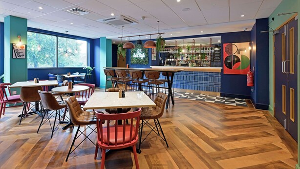 A restaurant with wooden flooring, multiple tables and chairs, a bar area, and large windows. - Travelodge Luton (Luton)