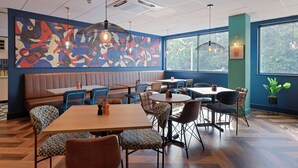 A restaurant interior with booths, tables, and chairs, featuring a large abstract mural on the wall and large windows. - Travelodge Luton (Luton)