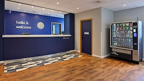 A reception area with a blue wall, a clock, a reception desk, and a vending machine. - Travelodge Luton (Luton)
