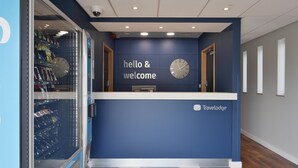 Un comptoir d’accueil avec une horloge et une pancarte indiquant « Bonjour et bienvenue », des distributeurs automatiques et le logo de Travelodge.