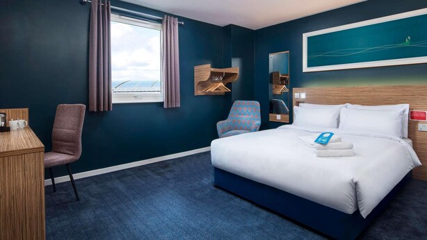 Une chambre d’hôtel avec un grand lit, un bureau avec une chaise, une fenêtre avec des rideaux, un miroir fixé au mur et un tableau au-dessus du lit.