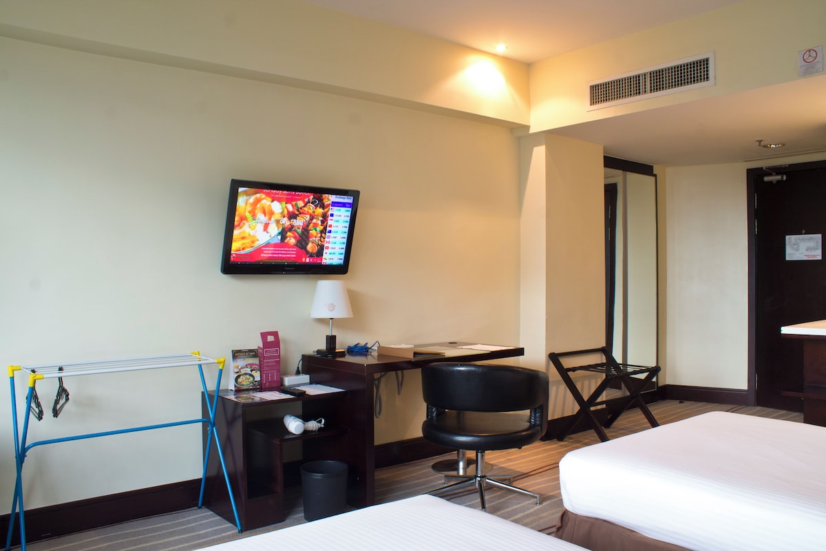 A hotel room with a bed, a desk with a computer, a chair, a TV on the wall, and a clothes drying rack.