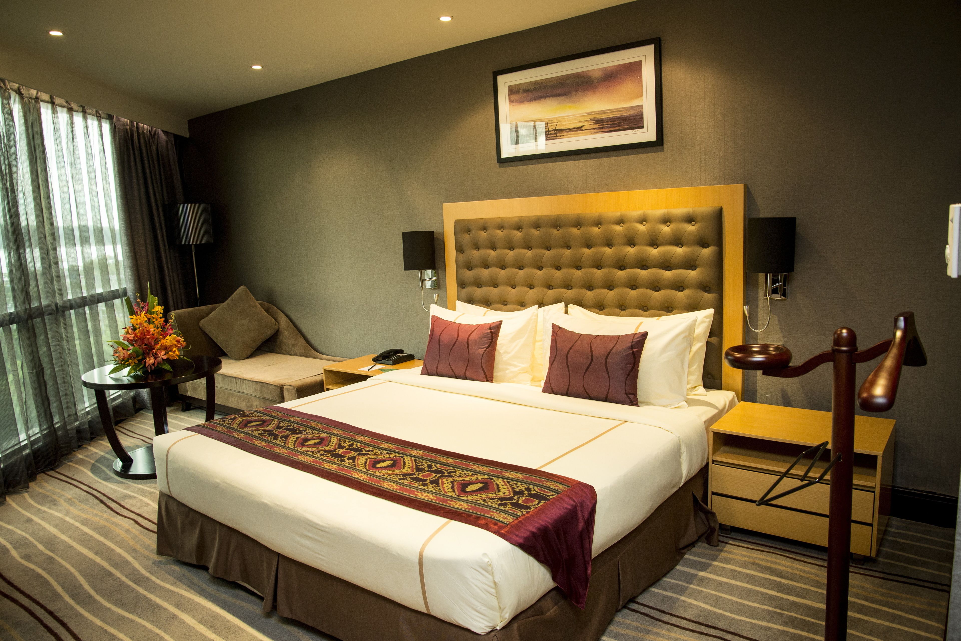 A hotel room with a large bed, a bedside table, a lamp, a chair, and a view of the outdoors through the window.