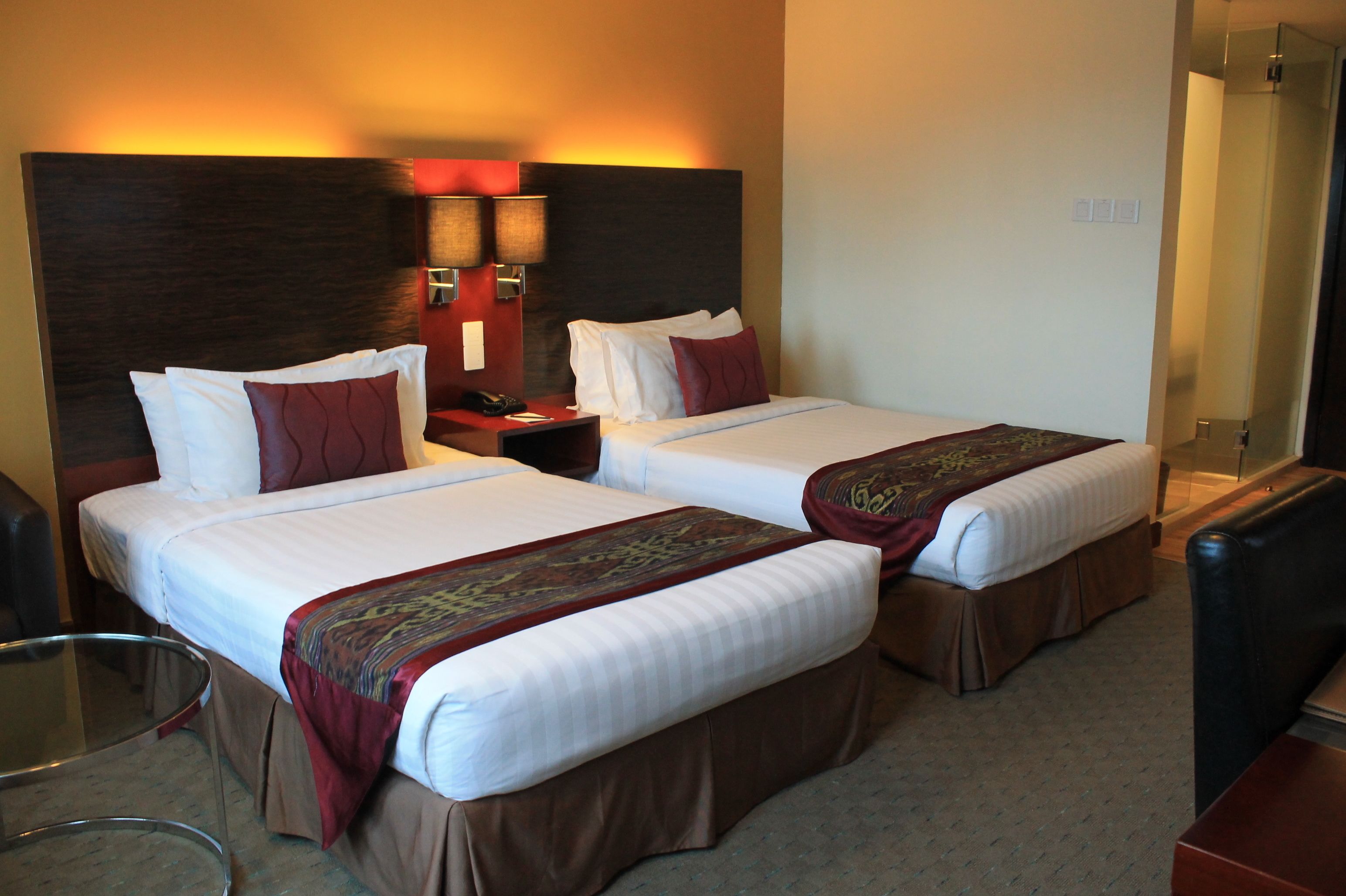 A hotel room with two beds, a nightstand, a lamp, and a glass table.