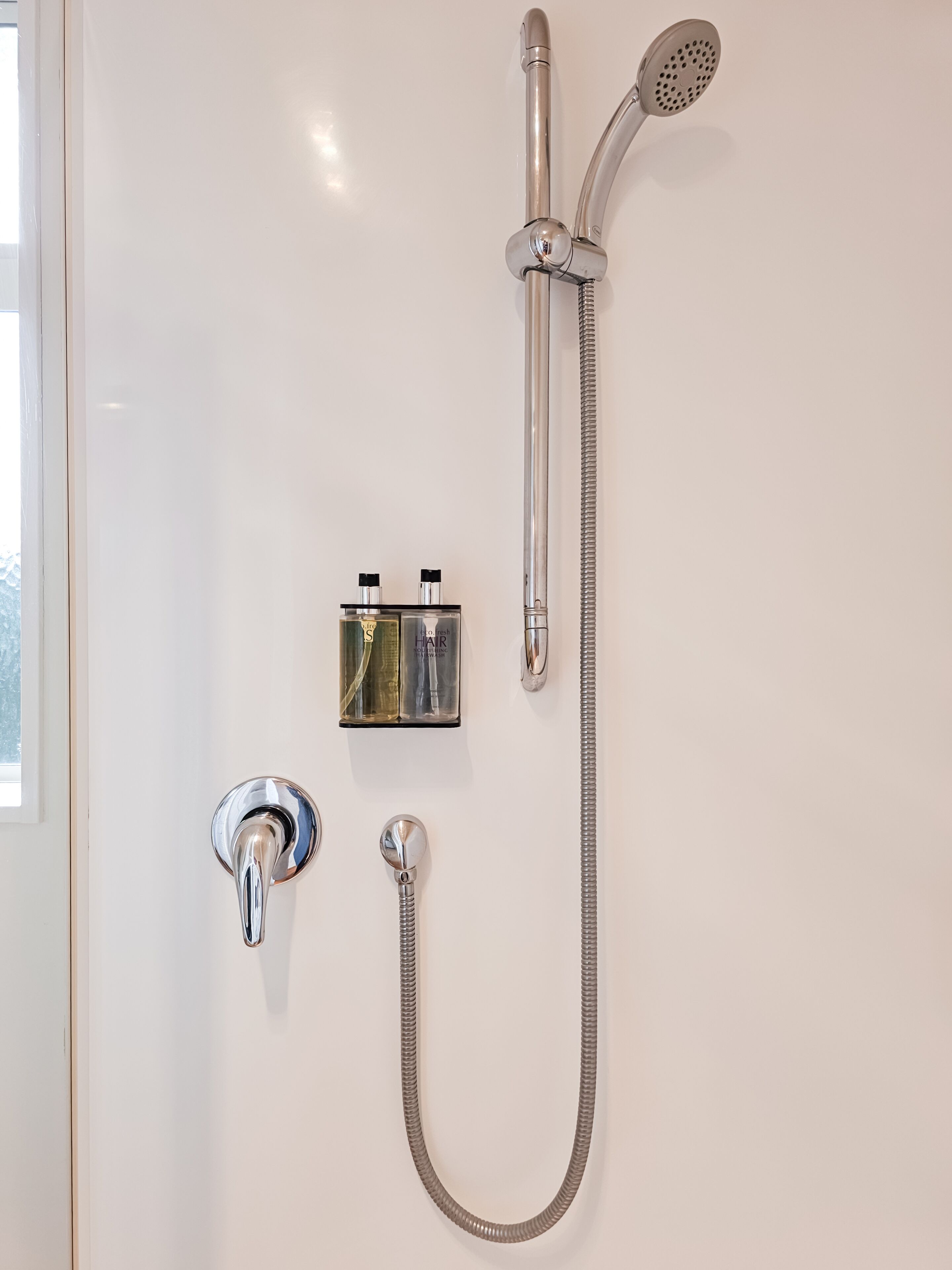 Un baño con ducha, cabezal de ducha de mano, grifo de pared y una botella de champú.
