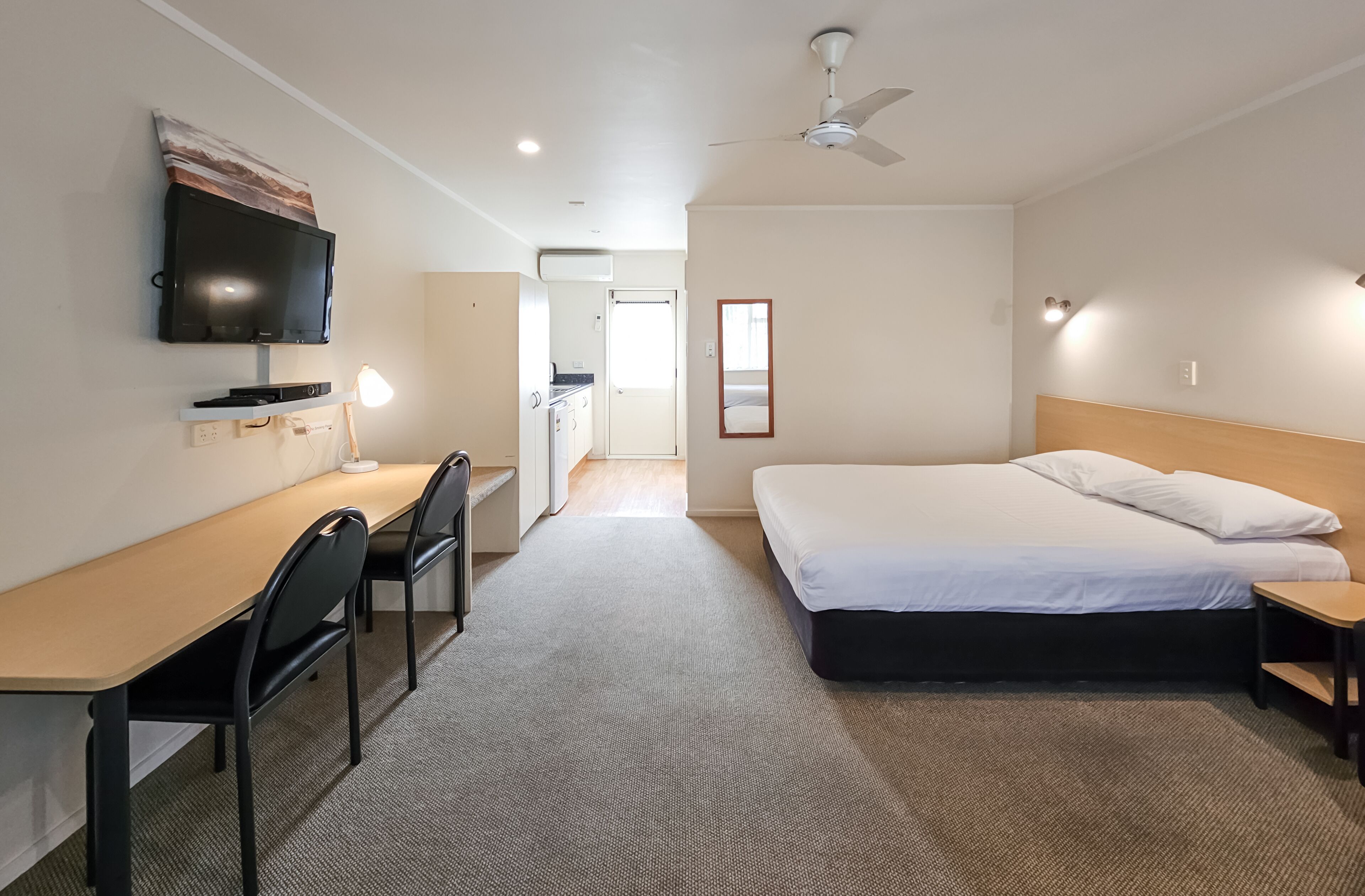 A hotel room with a bed, desk, chairs, a TV, and a ceiling fan.