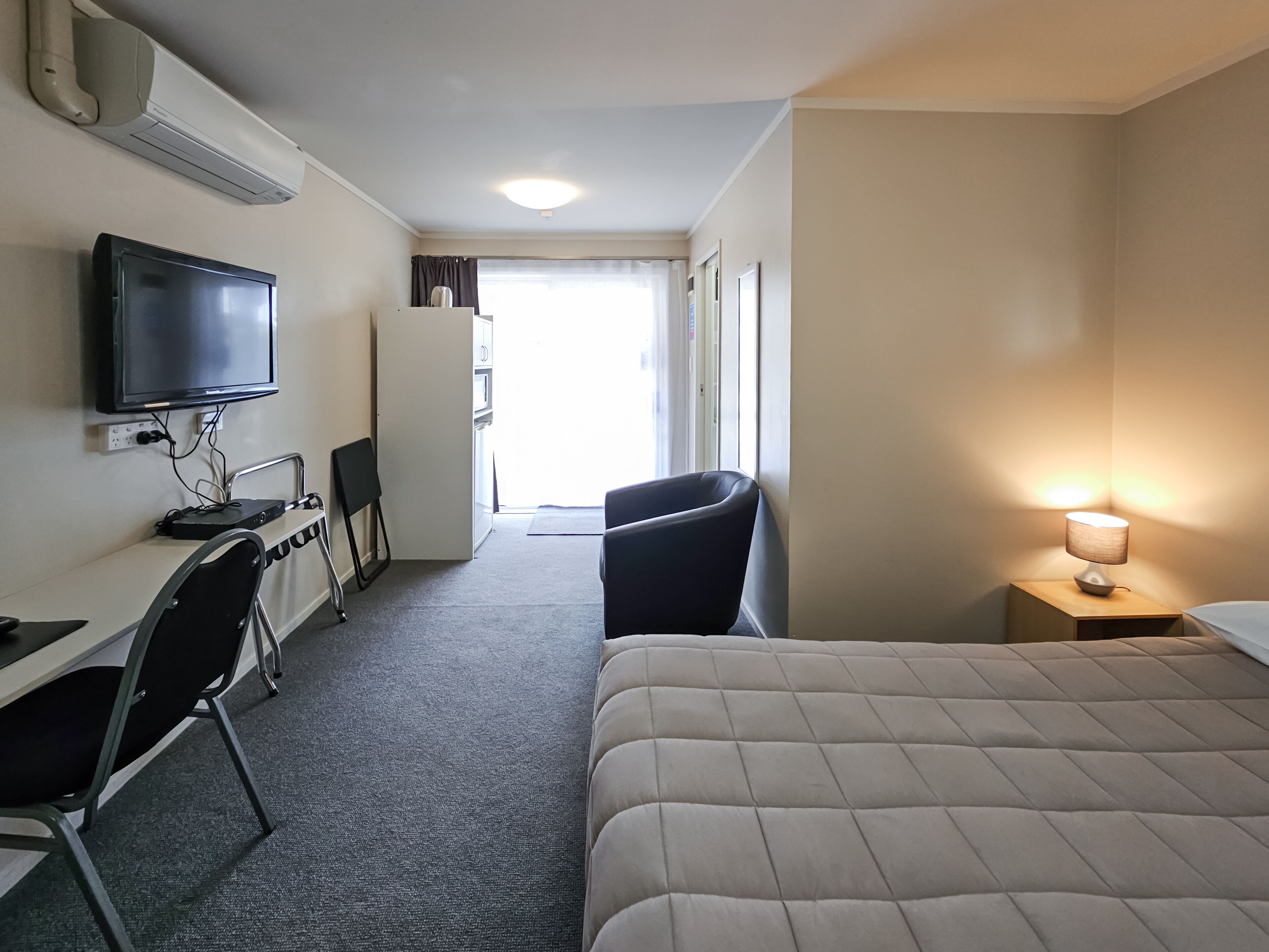 A hotel room with a bed, a desk with a computer, a chair, a TV, and a bedside lamp.
