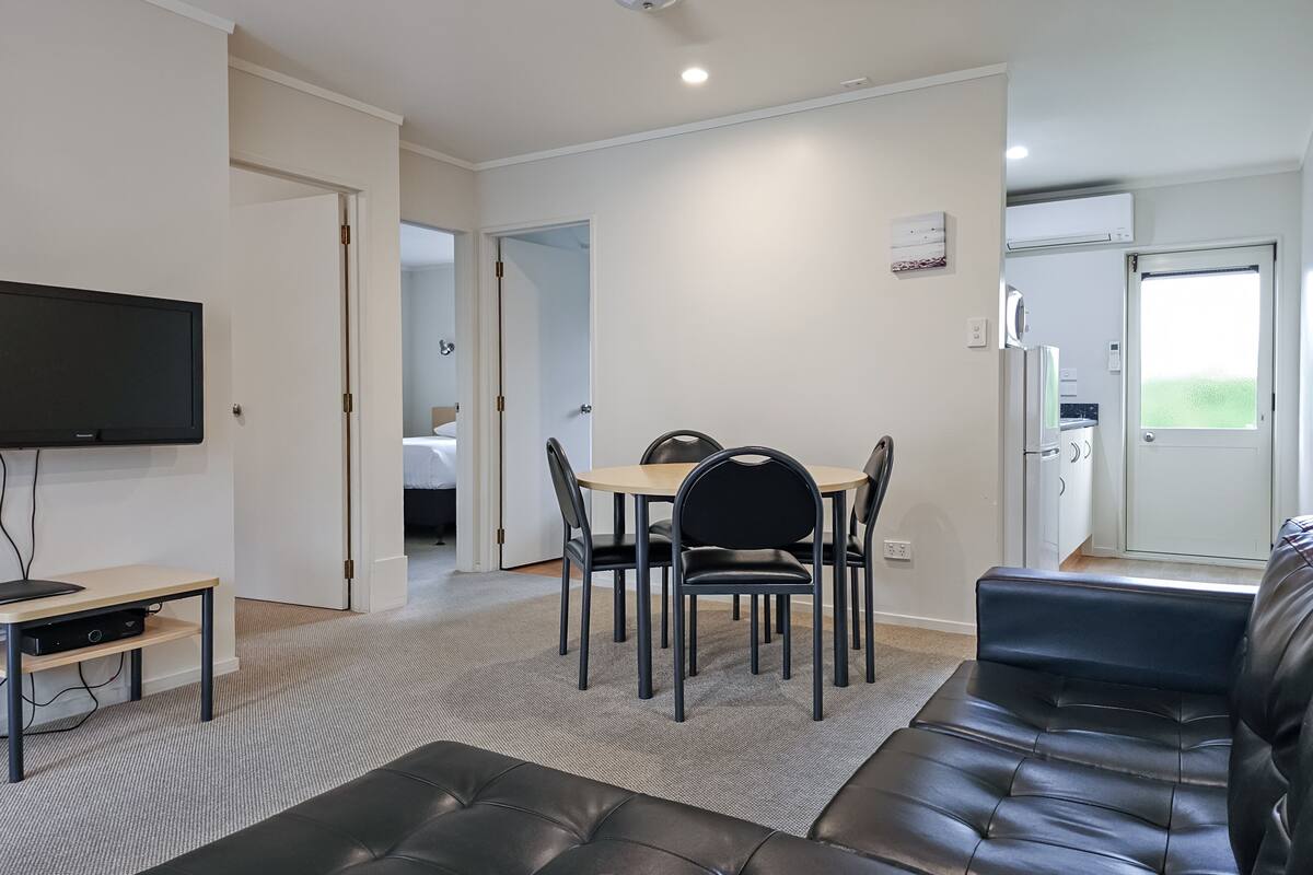 A modern living room with a black leather sofa, a dining table with chairs, a flat-screen TV, and a kitchen area visible through an open door.
