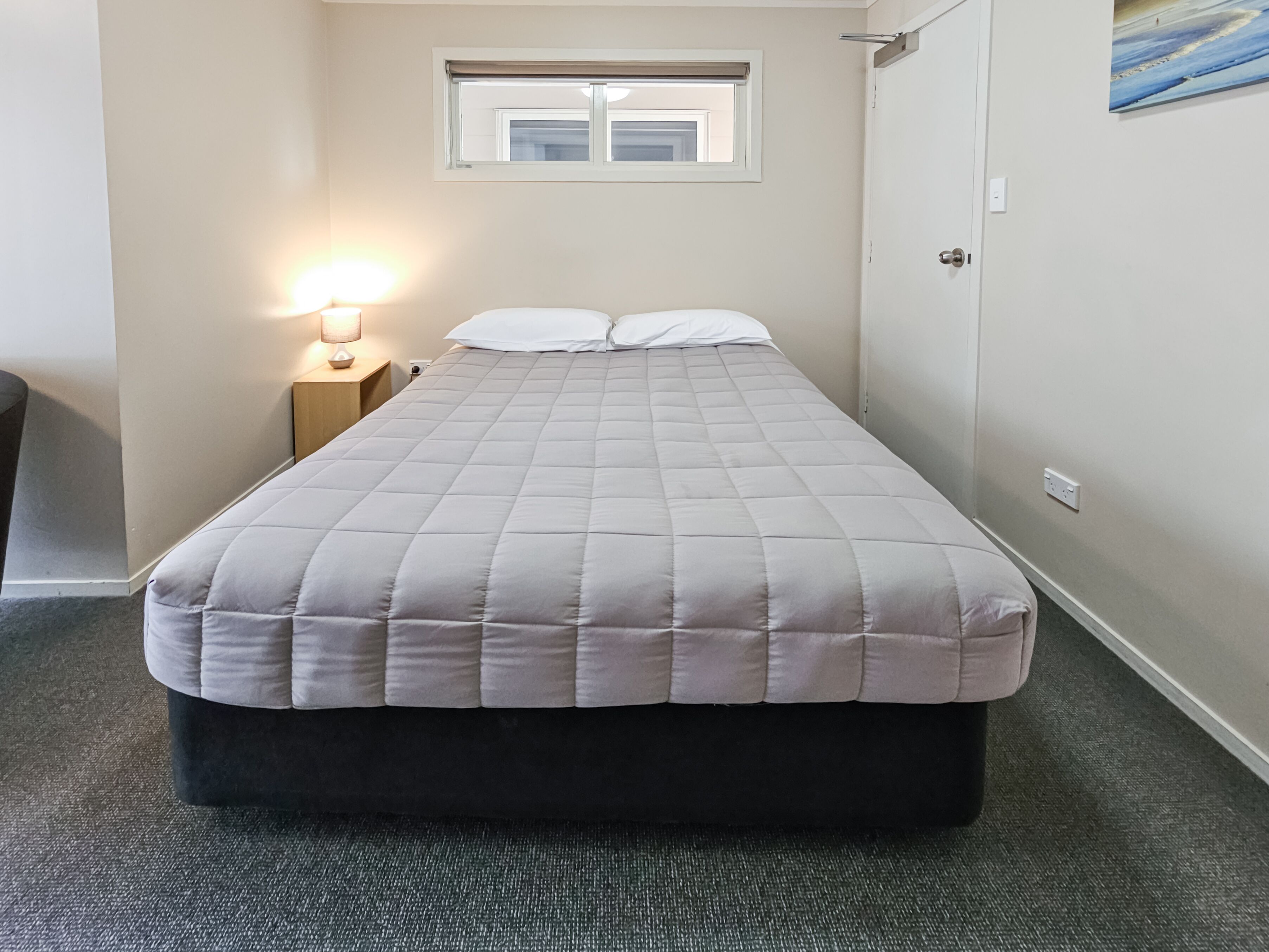 Una cama individual con colcha gris, una lámpara de noche y una ventana con persianas.