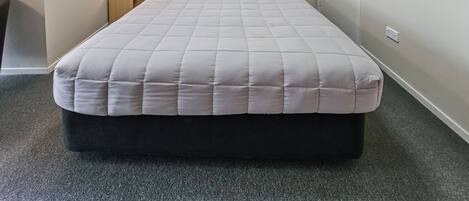 Una cama individual con colcha gris, una lámpara de noche y una ventana con persianas.