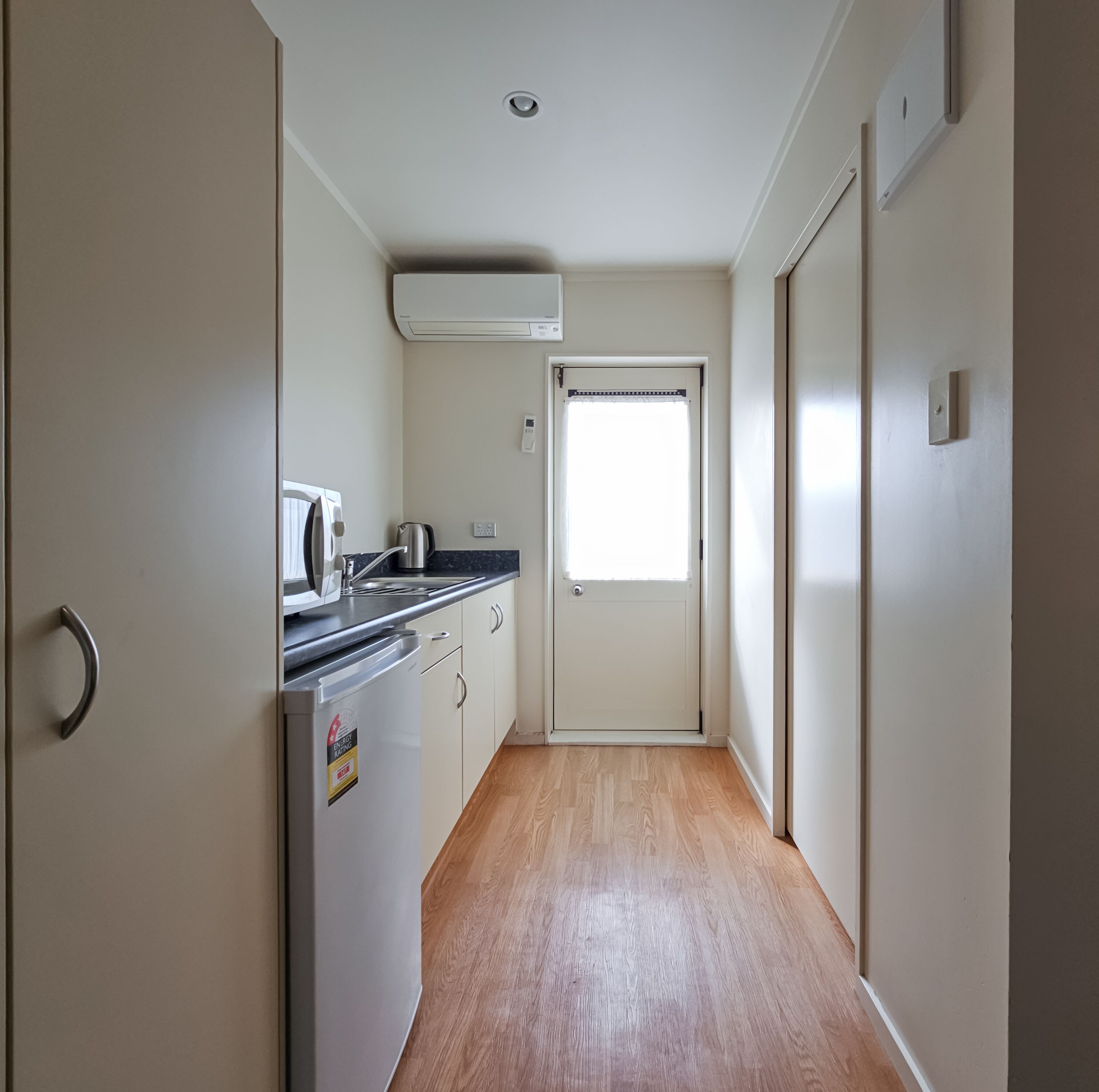 Un estrecho pasillo con una zona de cocina a la izquierda, que incluye un refrigerador, estufa y lavabo. Se ve una ventana con persianas en el fondo.