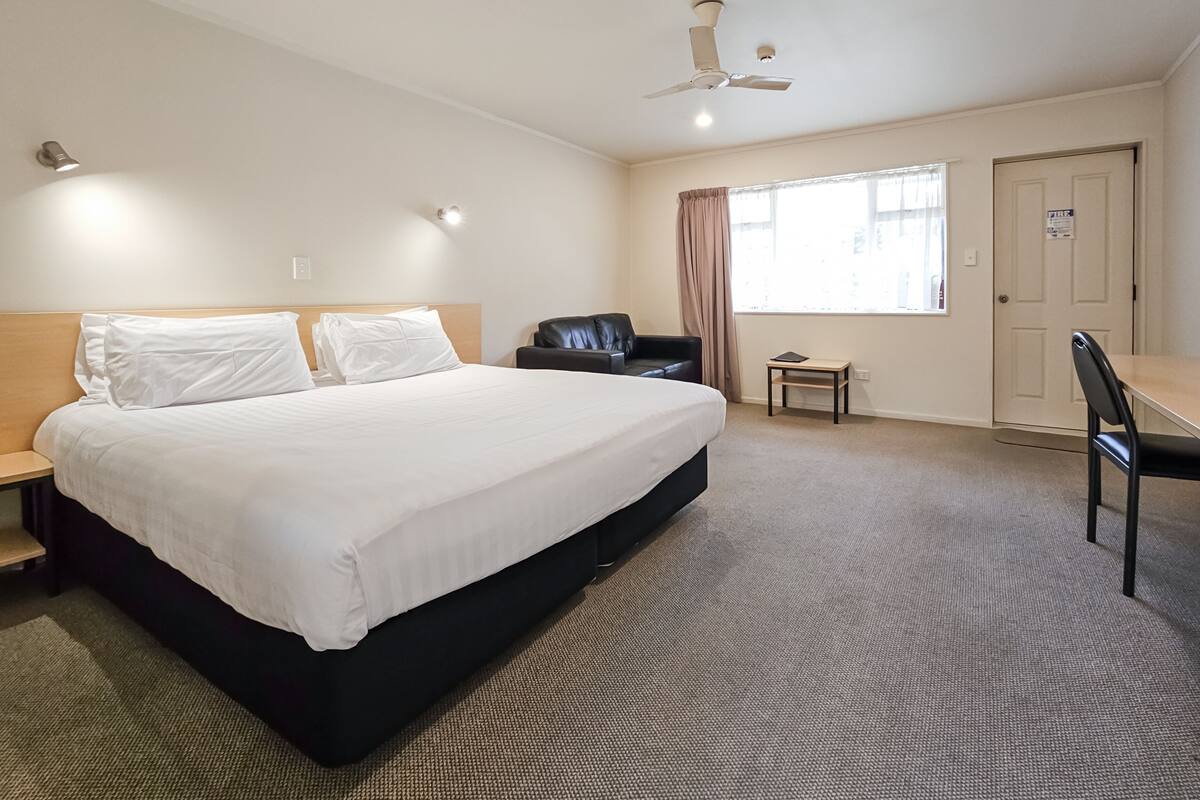 A hotel room with a large bed, a desk, a chair, a ceiling fan, and a window with curtains.