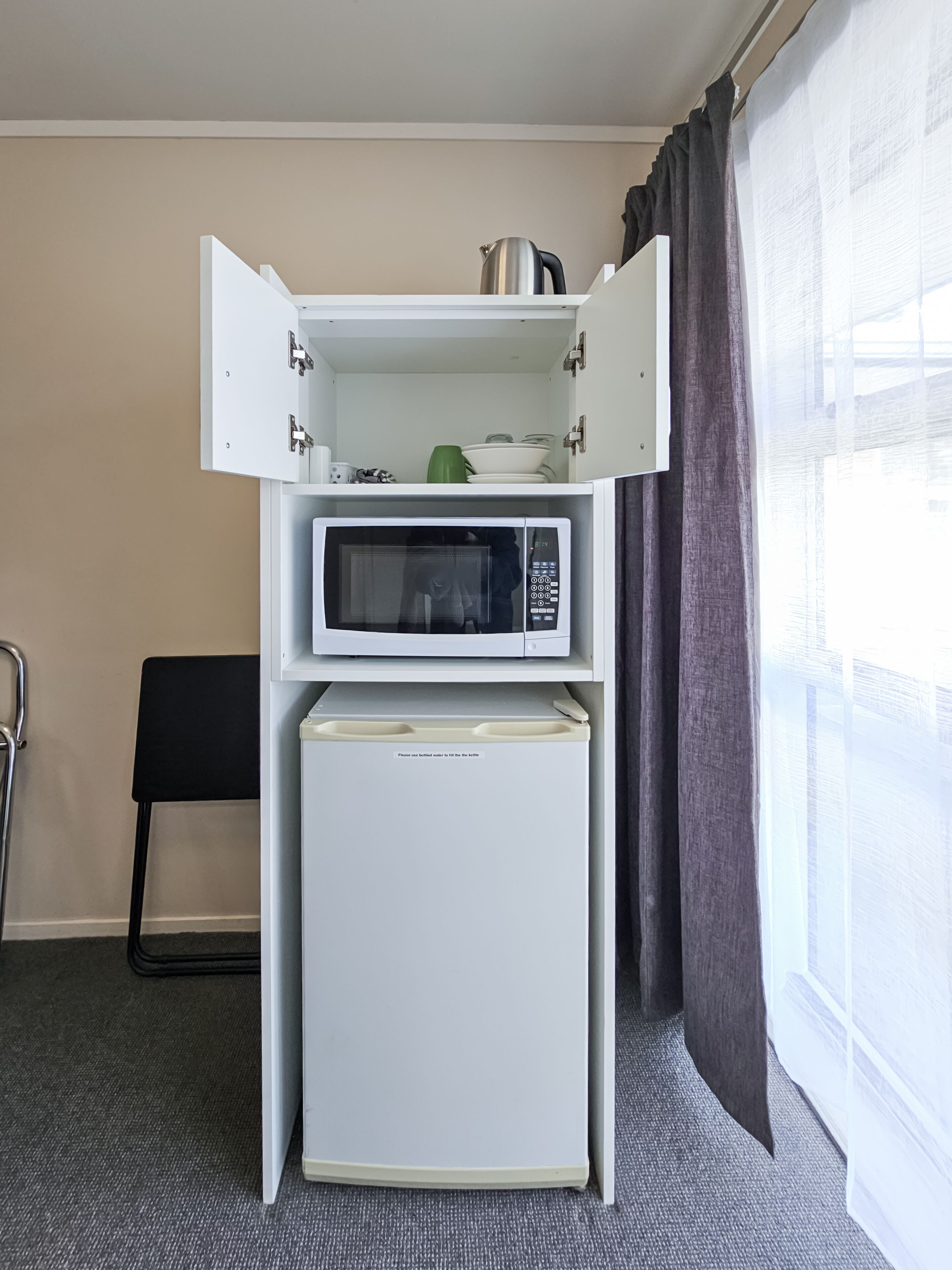A compact kitchenette with a microwave and refrigerator, a chair, and a window with curtains.
