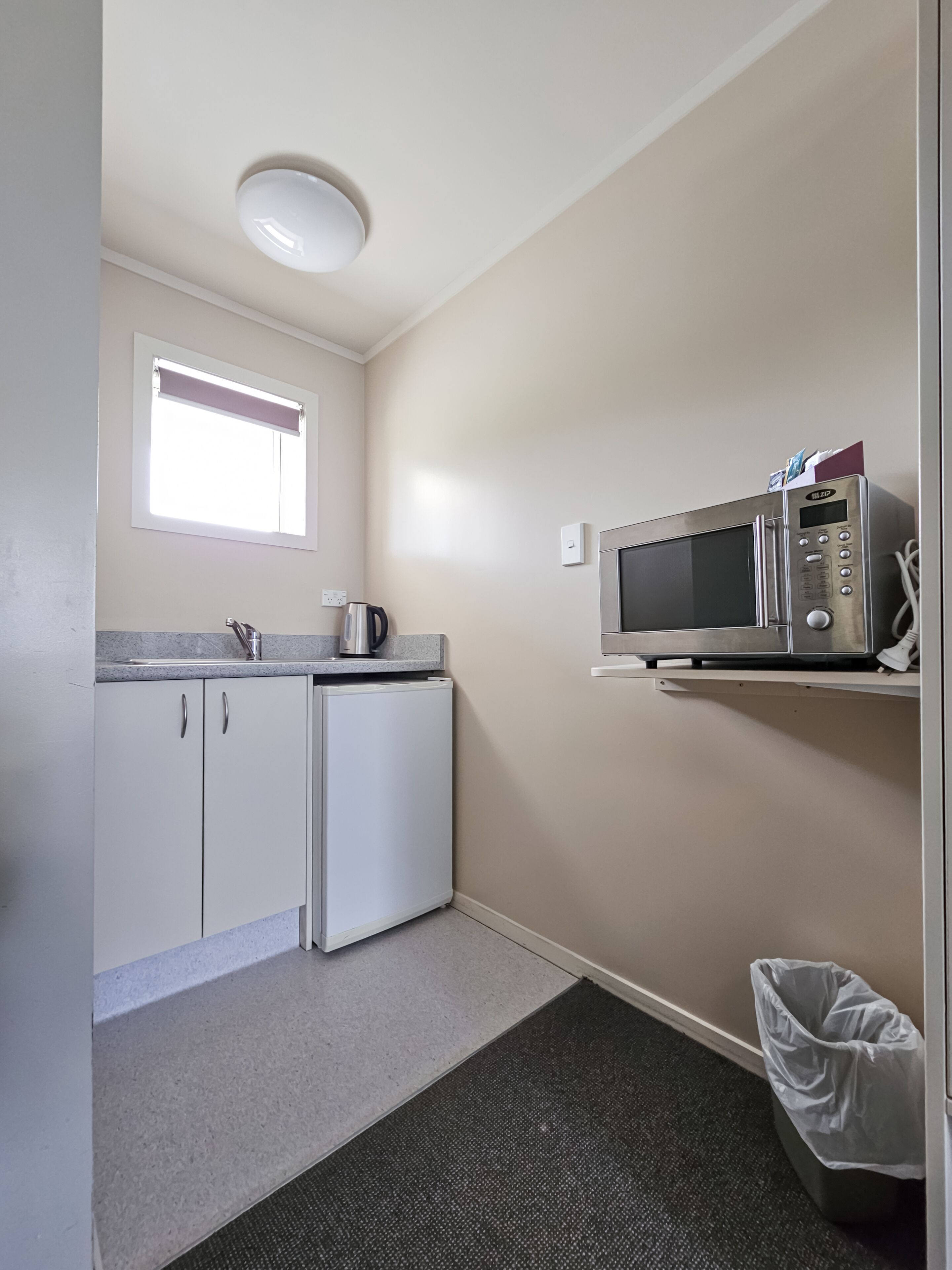 Una cocina compacta con armarios blancos, microondas integrado, hervidor y una ventana pequeña.
