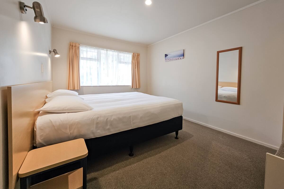 A hotel room with a bed, a desk, a chair, a window with curtains, a wall-mounted mirror, and a picture.