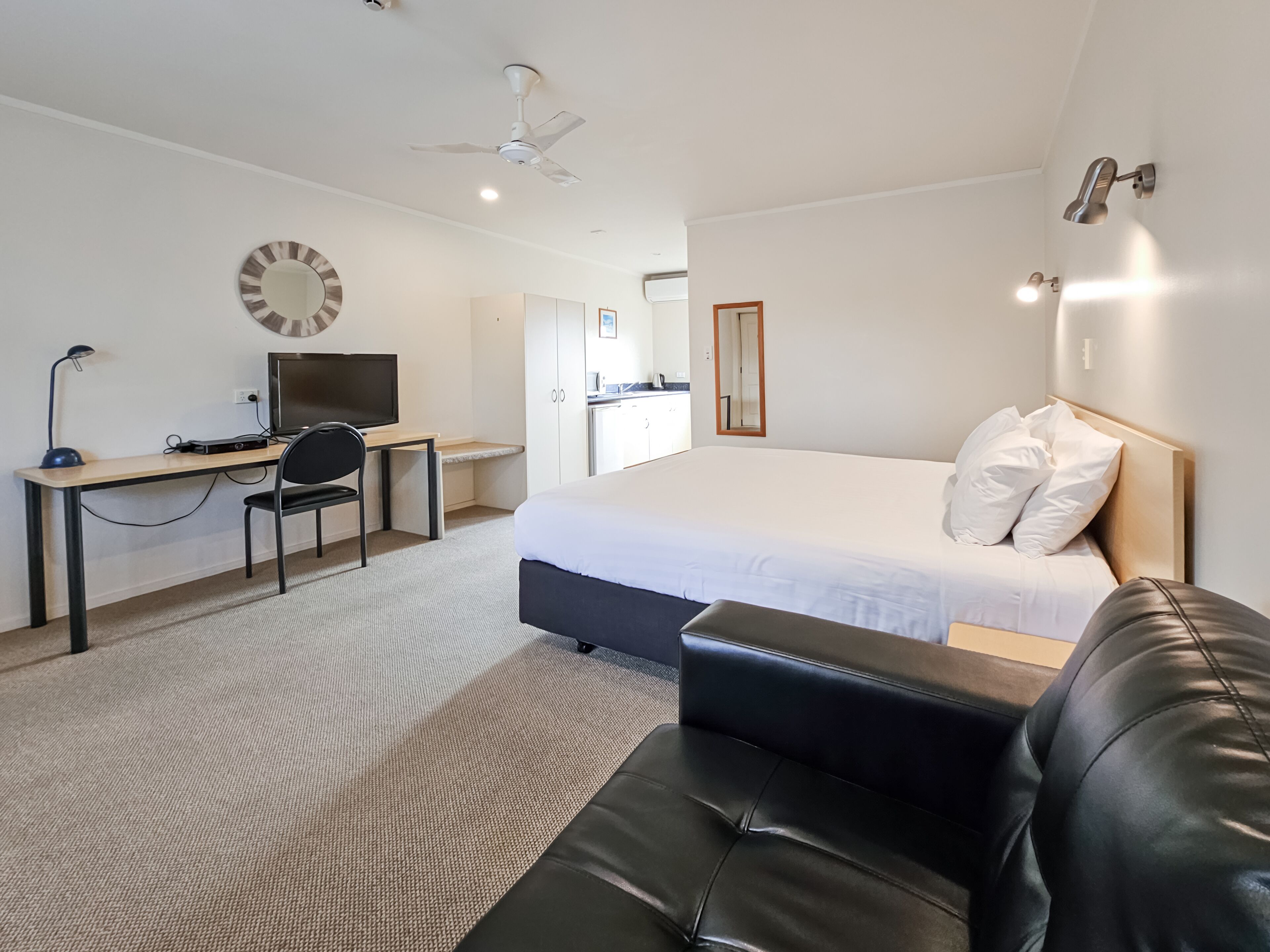 Quarto de hotel com cama, escrivaninha com cadeira, televisão, ventoinha no teto e uma pequena cozinha compacta ao fundo.