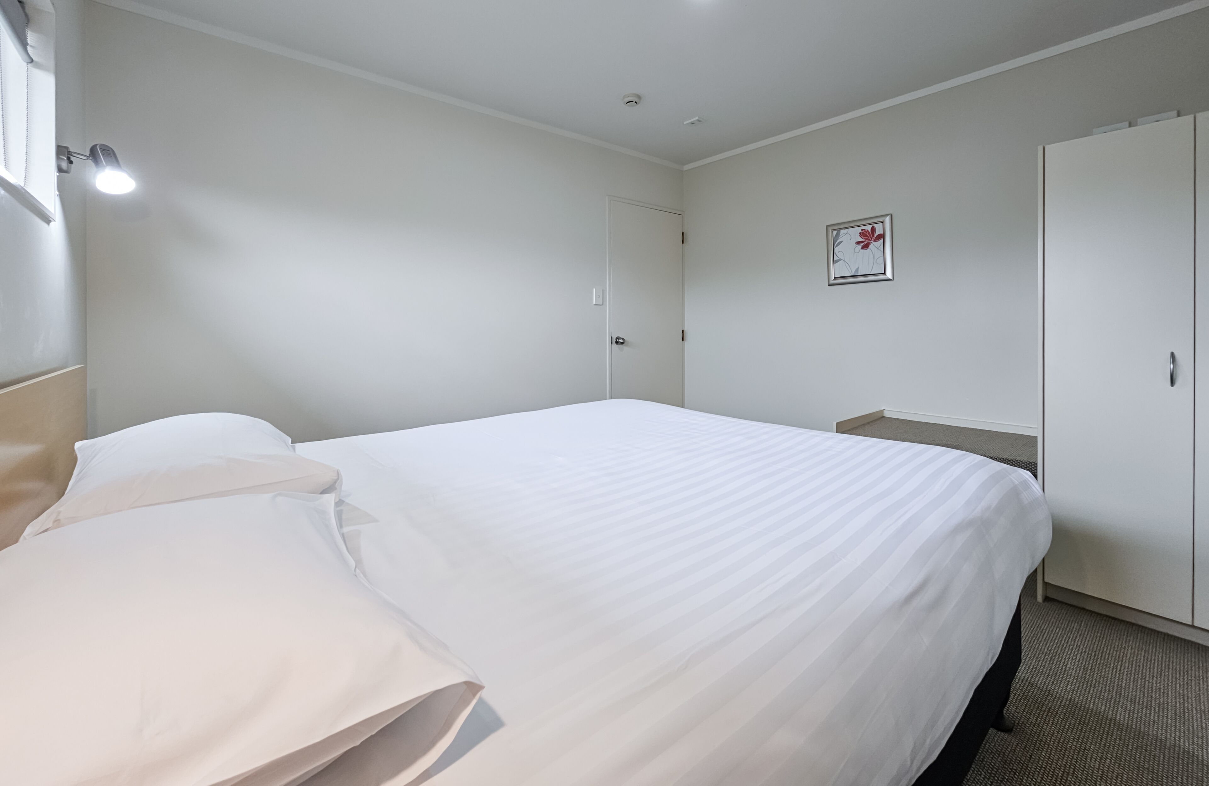 A hotel room with a large bed, a wardrobe, a bedside table, and a wall-mounted light.