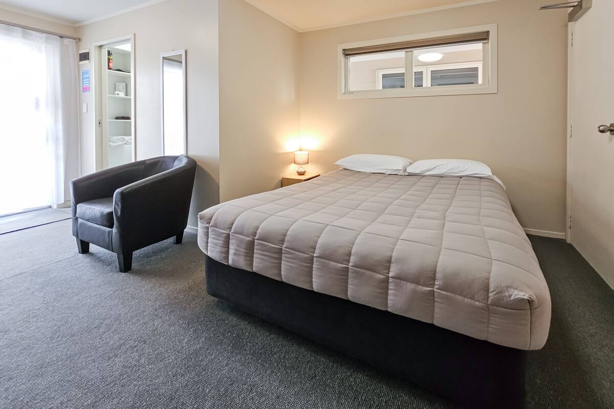 A hotel room with a bed, a chair, a nightstand with a lamp, a window, and a door.