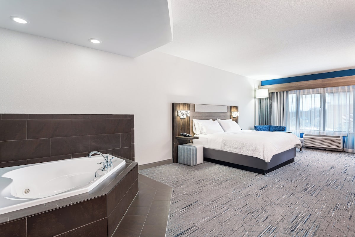 A modern hotel room with a large bathtub, a neatly made bed, and a view of the outdoors through large windows.