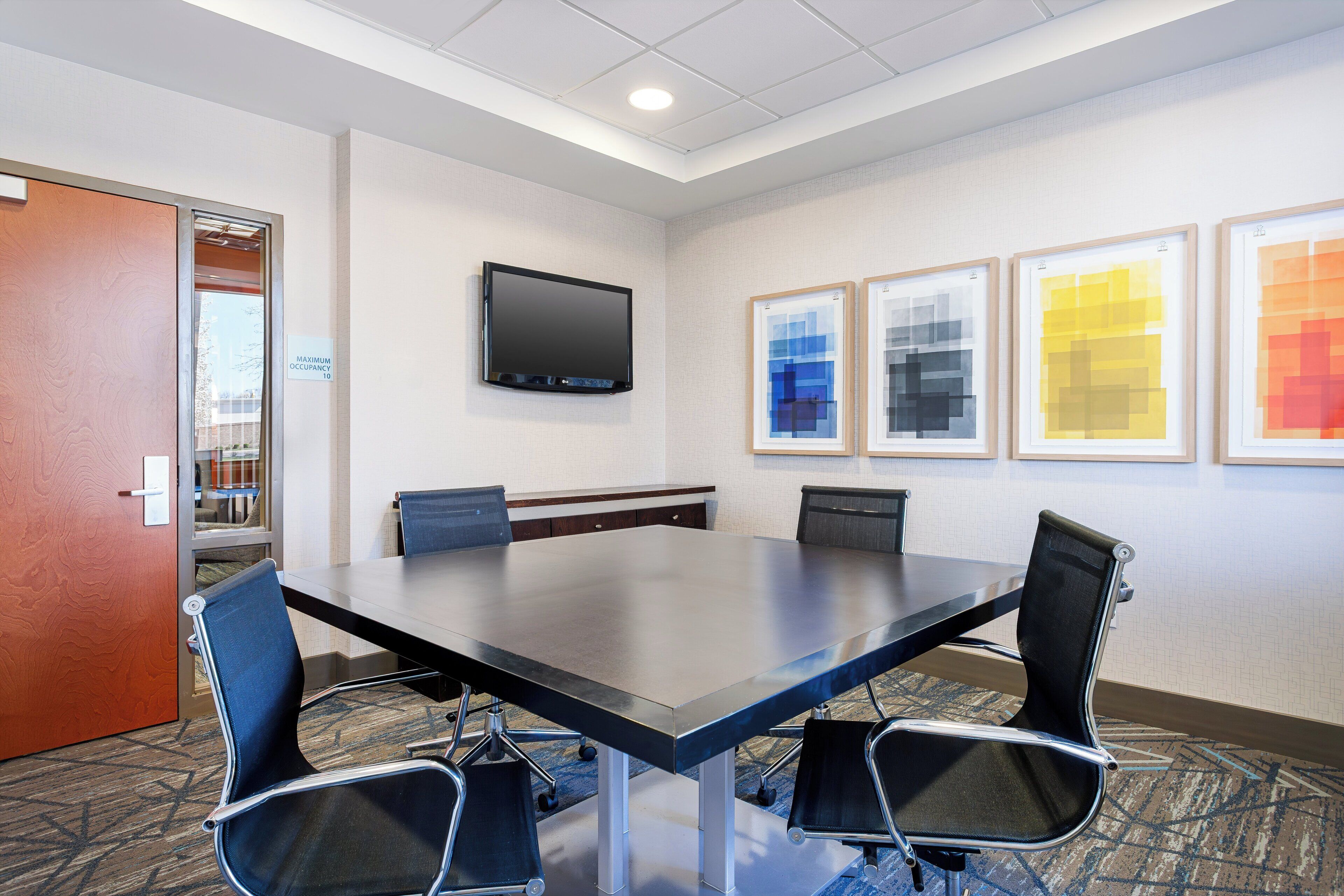 A conference room with a long table, four chairs, a flat-screen TV, and three framed artworks on the wall.