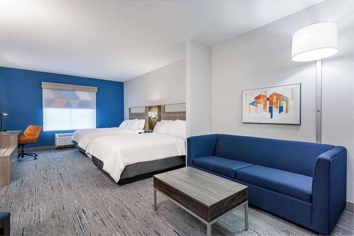 A hotel room with a bed, a blue sofa, a wooden coffee table, a desk with a chair, and a painting on the wall.