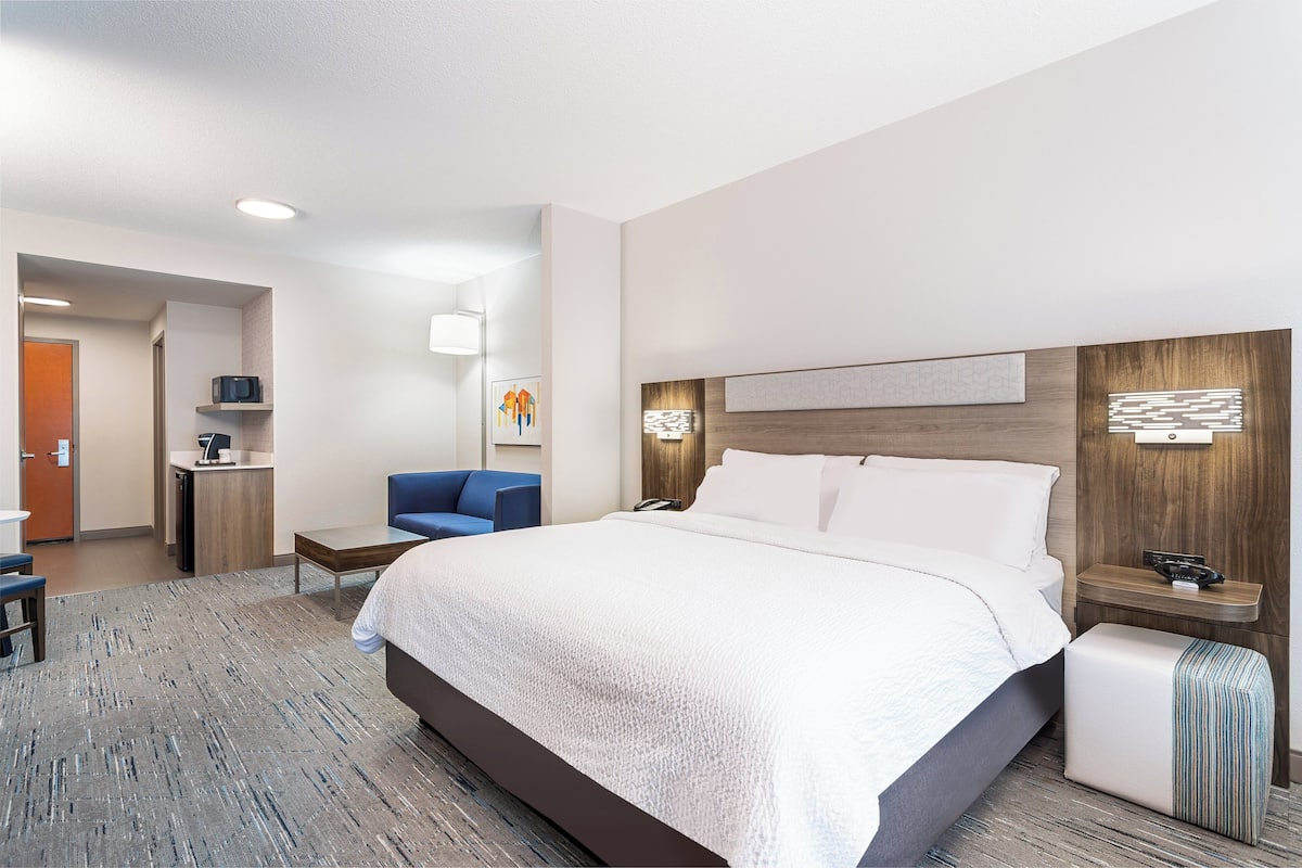A hotel room with a large bed, a desk with a chair, a small kitchenette, and a bathroom.