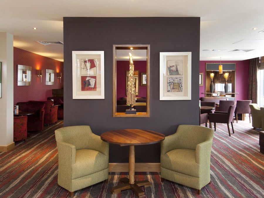 A hotel lobby with a round wooden table, two green armchairs, a red sofa, and framed artwork on the walls.