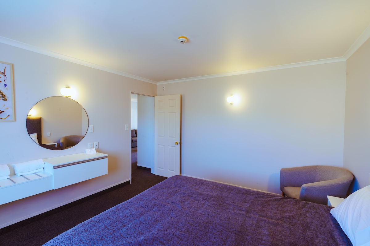 Quarto de hotel com uma cama, um espelho redondo, uma mesinha de cabeceira branca, uma cadeira e uma porta que dá para outro quarto.