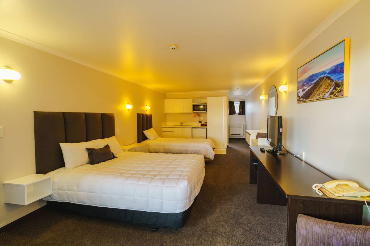 Quarto de hotel com duas camas, uma escrivaninha, televisão e cozinha ao fundo.