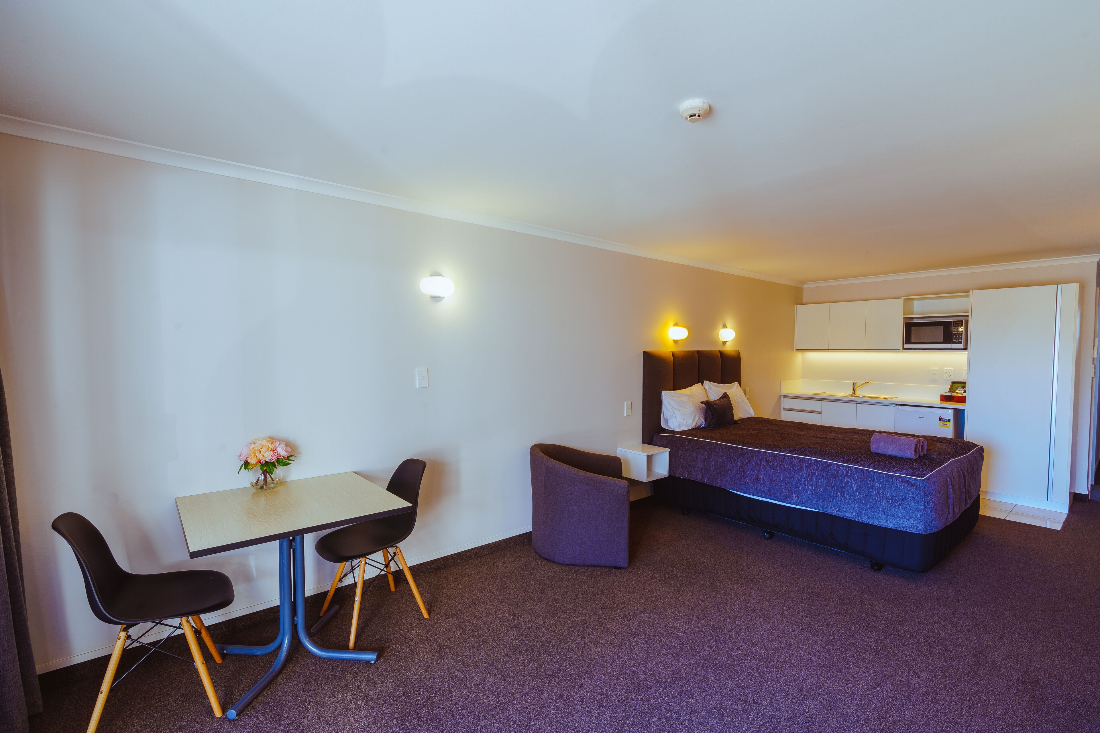 Une chambre d’hôtel compacte comprenant un lit, une petite kitchenette, un coin repas avec une table et des chaises, et un seul luminaire fixé au mur.