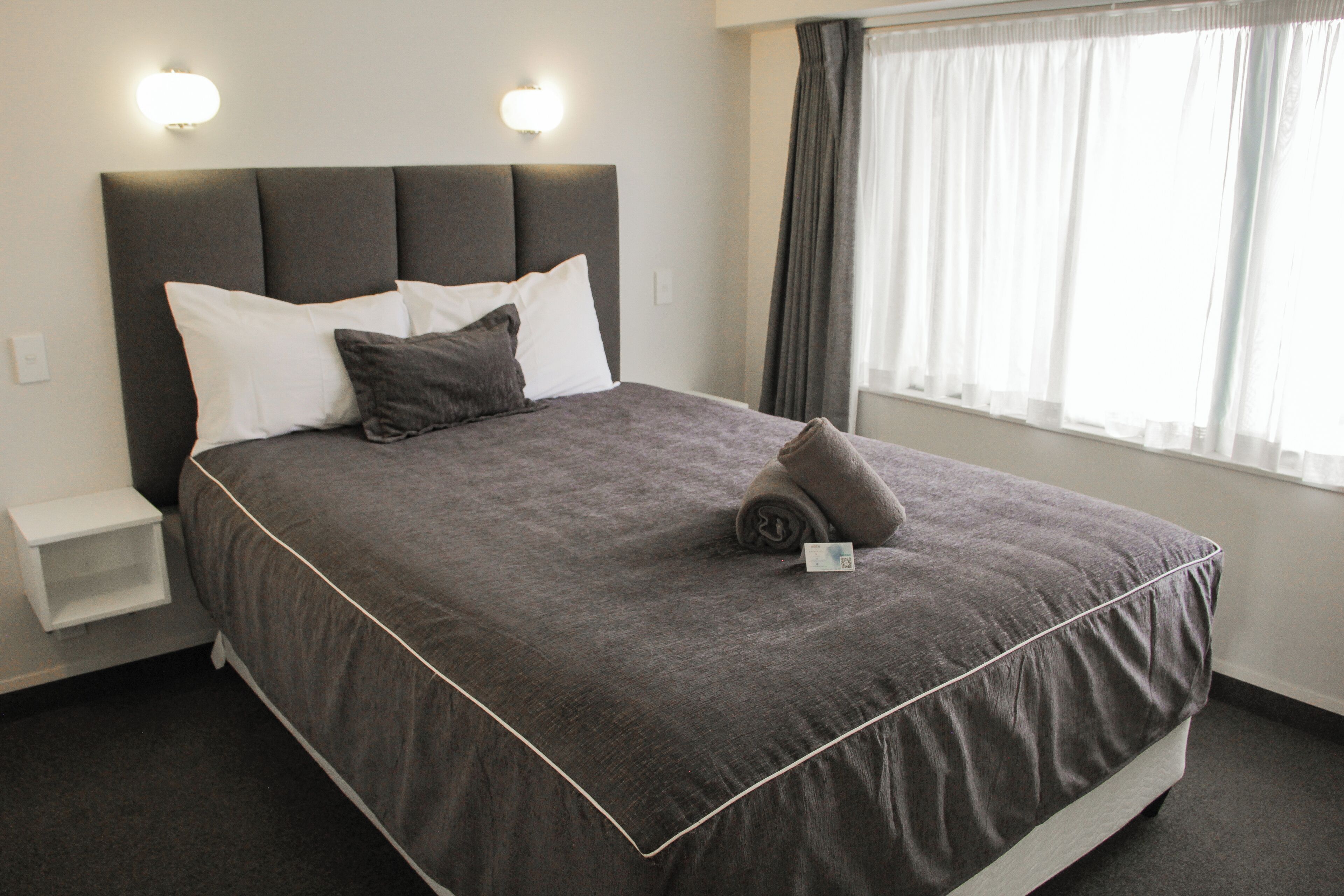 A neatly made bed with a grey bedspread and pillows, a bedside table, and a window with curtains.