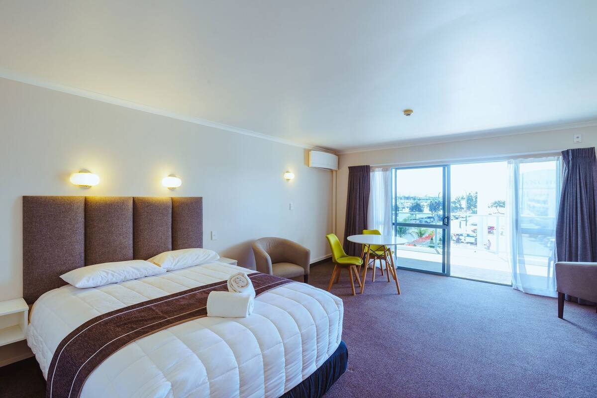 Quarto de hotel com uma cama grande, uma escrivaninha com duas cadeiras, uma janela com cortinas e vista para o exterior.