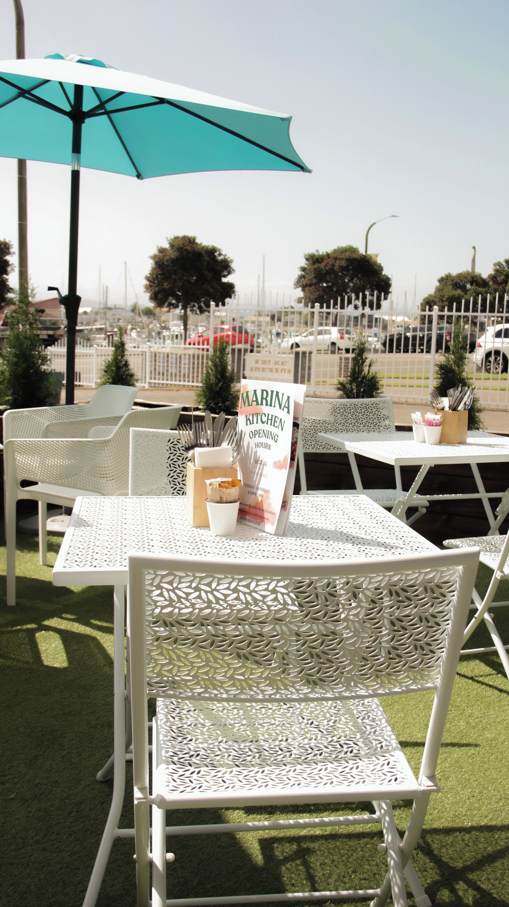 Outdoor seating with white metal chairs and tables, a sign for 'Marina Kitchen Opening Hours', and a blue umbrella.