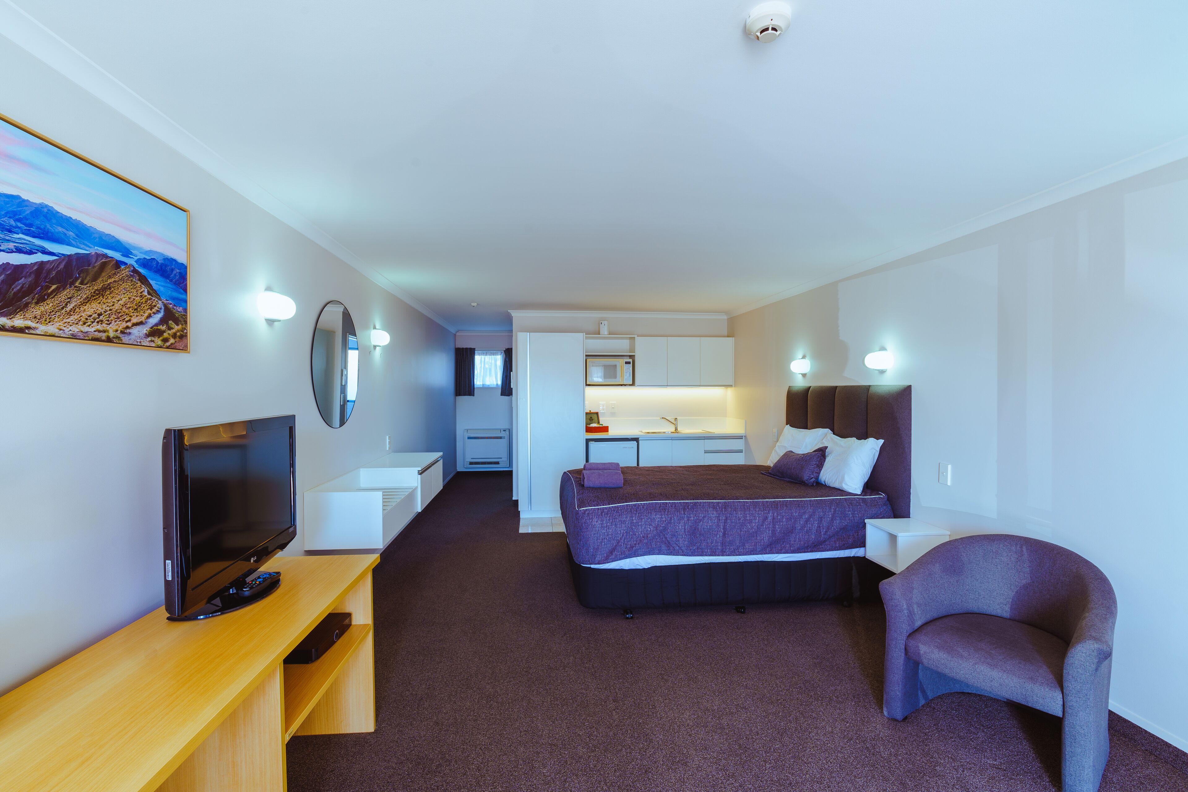 A modern hotel room with a bed, a TV, a desk, a chair, and a bathroom visible in the background.