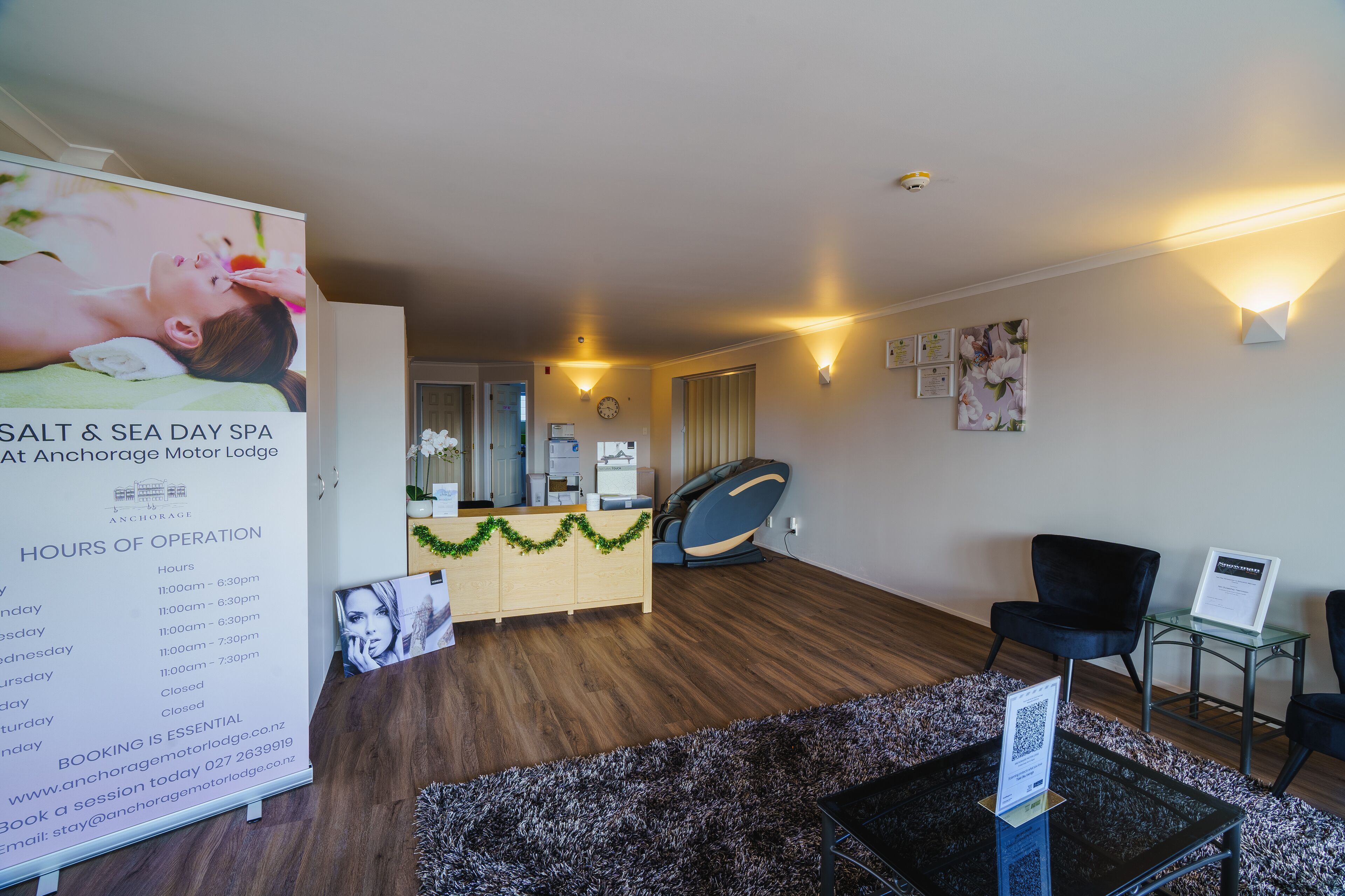 Zona de recepción de un spa con suelo de madera, un mostrador de recepción y un reloj fijado a la pared. Hay dos sillas y una pequeña mesa con un cartel.